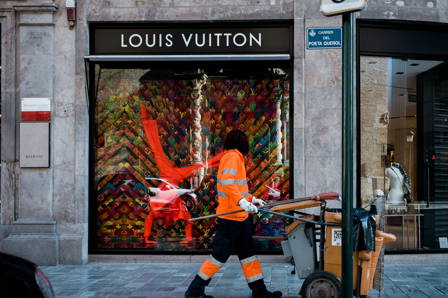 Street cleaner pushes a cart past a luxury boutique storefront.