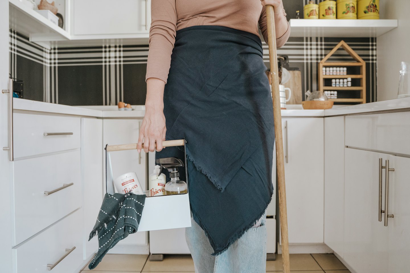 A person in an apron holds cleaning supplies in a kitchen.